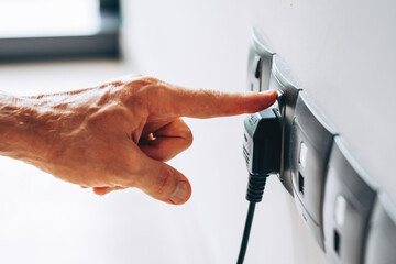 Hand plugging a black electrical cord into one of several sockets on a white wall. Close view with focus on safe energy use. Energy use, household electricity, safe connection concept.