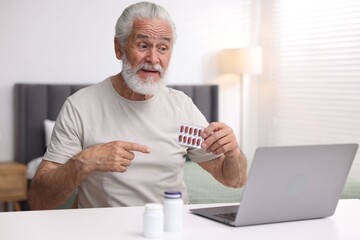 Senior man with pills having online consultation with doctor via laptop at table indoors