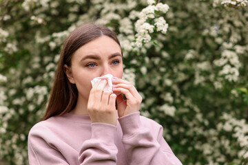 Woman with paper tissue suffering from seasonal pollen allergy near blossoming shrub on spring day,...