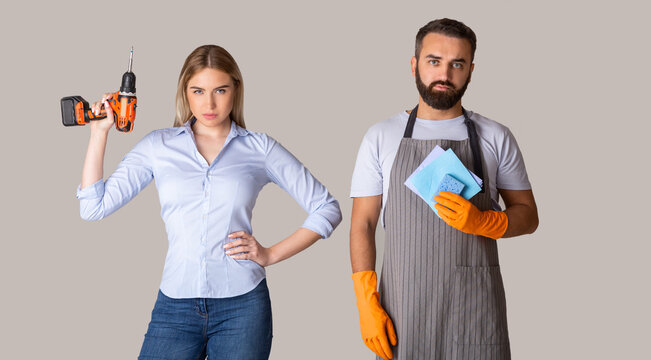 Gender stereotypes and non female profession. Serious man in apron and rubber gloves holding sponges and woman holding a drill on gray background, studio shot