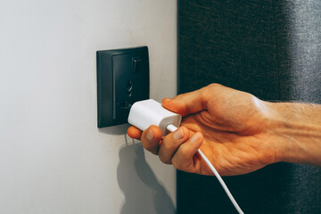 Hand inserting a white charging adapter into a wall socket. Close-up view inside a modern interior. Technology, charging, modern lifestyle concept.