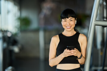 Smiling Woman in Gym Wearing Black Outfit While Using Smartphone and Earbuds