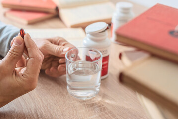 Man going to take pill from headache drinking clear water from glass. Preparing to take dietary supplement, man s hand with capsule, surrounded by books and water.