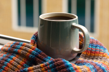 A mug of hot coffee or tea wrapped in a scarf against the backdrop of a city street. The onset of autumn or winter. Coffee and tea on the balcony windowsill.