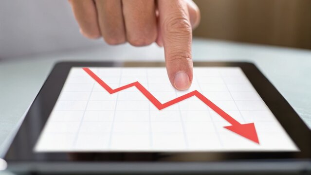A finger points at a downward trend on a tablet screen, showcasing a financial decline represented by a red arrow.