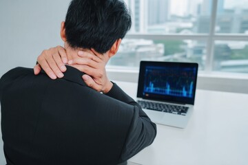 Young businessman suffering from neck pain while working on laptop in modern office environment. discomfort is evident as he holds his neck, reflecting stress and fatigue
