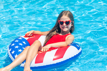 Happy smiling little girl 7-8 years old in red swimsuit and sunglasses relaxing in outdoor swimming pool on lifebuoy decorated with American flag print. Vacation in USA concept. 4th of July pool party