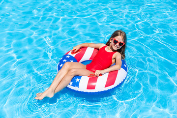 Happy smiling little girl 7-8 years old in red swimsuit and sunglasses lying on lifebuoy decorated with American flag print on blue water background. Vacation in USA concept. 4th of July pool party