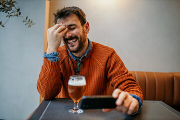 Cheerful man laughing while reading smartphone and drinking beer in pub