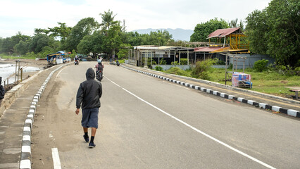 People walk and ride motorcycles along a coastal road with seaside stalls, green trees, and distant mountains under a cloudy sky in a small-town atmosphere.