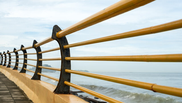 Closeup of yellow safety rail with modern black support on a seaside promenade, captured in a linear perspective. Calm ocean waves and soft cloudy sky in the background.