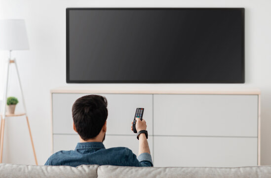 Man watching television, turning on plasma flatscreen TV-set, pointing remote control at empty TV screen on white wall. Guy switching channels at home, back view. Mockup