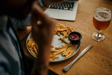 Businessman eating french fries with ketchup and drinking beer at his desk