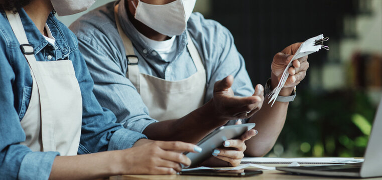 Finance, small business, COVID-19 pandemic. Sad millennial african american couple owners of small business in protective masks work with laptop and smartphone and accounts in cafe, cropped, panorama - Powered by Adobe