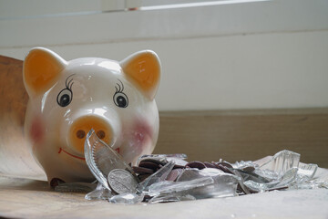 Broken piggy bank lies wooden surface, surrounded by scattered coins and shards of glass,...