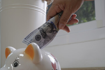 Hand placing stack of dollar bills into white piggy bank, symbolizing saving money and financial planning. scene is set indoors with natural light coming through window, creating calm atmosphere