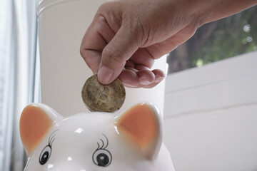 Hand places coin into piggy bank, symbolizing saving and financial growth. scene is set indoors with natural light, creating hopeful and secure atmosphere