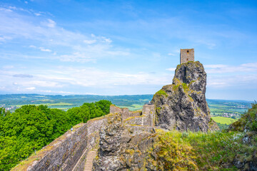 Visitors admire the historic Panna Tower perched atop a rocky outcrop at Trosky Castle Ruins in the scenic Bohemian Paradise, Czechia, surrounded by lush greenery and distant hills.