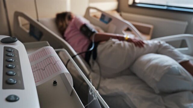 Close-up of a fetal monitor in a doctor's office, displaying fetal heart rate and uterine contraction data during a prenatal check-up with a blurred view of a pregnant woman lying in a hospital bed