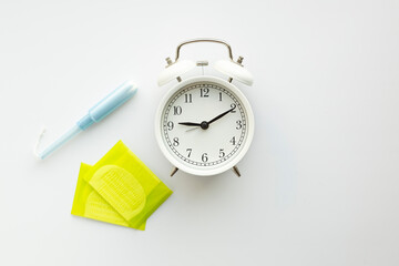 Flat lay of white alarm clock, tampon with applicator and pads on white.