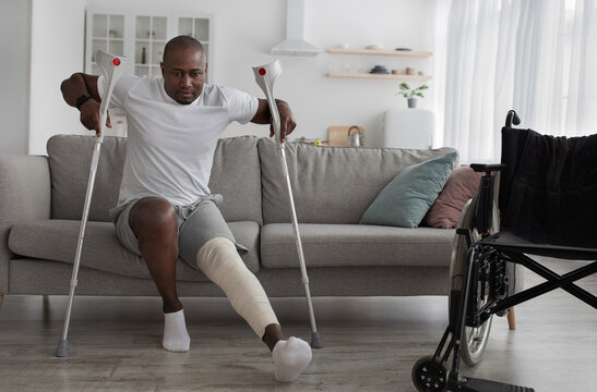 Bone fracture after incident, treatment at home, recovery. Black middle aged male with broken leg with plaster cast gets up from sofa with crutches in living room interior with wheelchair, empty space