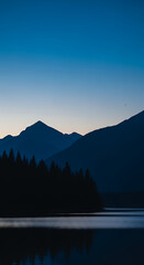 Tranquil mountain lake scene at dusk with silhouetted trees and layered peaks