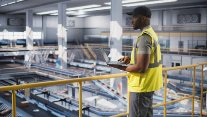 Young Black Man Standing on a Metal Platform, Using a Tablet Computer at a Post Office Logistics Center. African Specialist Working at a Sorting Facility, Configuring the Automation Process