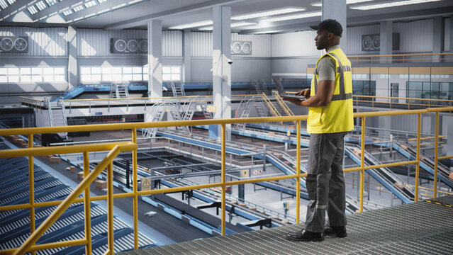 Black Employee Working at a Modern Logistics Center for an International Online Shopping Business. African Man Using a Laptop Computer to Optimize the Orders Before Deliveries - Powered by Adobe