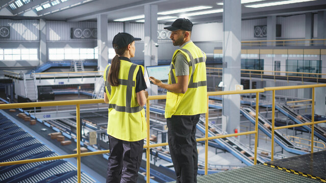 Employees Working at a Modern Logistics Center for an International Online Shopping Warehouse. Man Using a Laptop Computer and Talking to a Female Manager. Worker Passing Them with a Cardboard Box