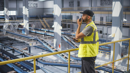 Logistics Manager Working at a Modern Logistics Center for an International Online Shopping Company. Multiethnic Man Using a Tablet Computer and a Two-Way Radio to Communicate with Colleagues