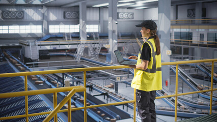 Female Working in a Modern Advanced Logistics Center with Automated Belt Conveyors for Sorting Packages. Young Woman in High Visibility Vest Using Laptop Computer in an Industrial Facility