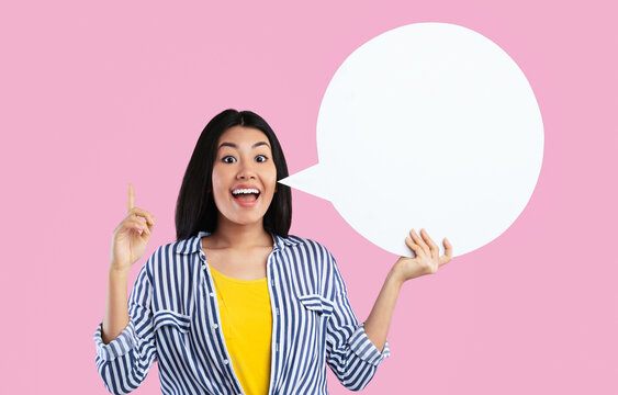 Inspired young asian woman having idea, holding blank speech bubble and pointing finger up, showing free place for your text, standing over pink background in studio, copy empty space