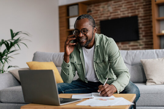Smiling busy mature african american guy manager in glasses and casual, have call by phone, works on laptop in living room interior. Business remotely, freelance, new normal with device and meeting - Powered by Adobe