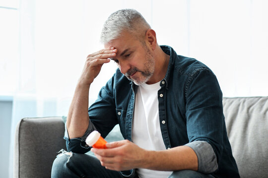 Senior man having headache, touching his temples while sitting on couch in living room, holding jar with pills. Grey-haired man suffering from migraine or tension problem, feeling sick