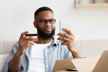 Quick shopping from the couch. African american guy with smartphone and credit card makes purchase