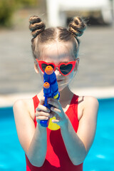 Laughing child girl in red swimsuit and sunglasses playing with water gun in outdoor swimming pool on summer day. Pool toys and water fun for family with children. Vacation and outdoor sport with kids