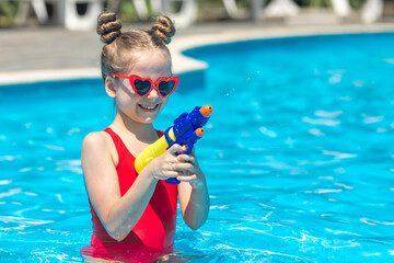 Laughing child girl in red swimsuit and sunglasses playing with water gun in outdoor swimming pool on summer day. Pool toys and water fun for family with children. Vacation and outdoor sport with kids