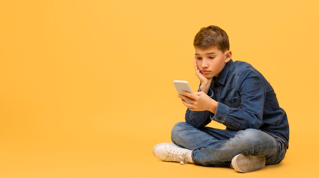 Bored teen boy using smartphone while sitting on floor in studio, upset male child looking at mobile phone screen, playing online games, feeling lonely, posing on yellow background, copy space - Powered by Adobe