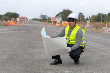 A male civil engineer He is carefully reviewing a large blueprint or construction plan, with a...
