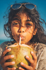 Young South Asian girl with tousled hair drinking coconut water under bright sky, close-up portrait filled with sunlight, ideal for vacation, summer and eco-themed content. happy, hydration, natural
