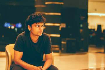 Pensive young Indian man in black t-shirt sits in a chair in a modern, softly lit Indian building...