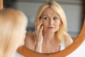 Skin aging concept. Beautiful aged woman looking at mirror at her wrinkles near eyes, checking...