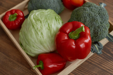 Fresh vegetables including iceberg lettuce, red bell peppers, broccoli and tomato arranged on wooden tray, showcasing vibrant produce for healthy cooking or food photography projects