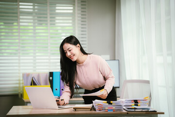 A cheerful businesswoman organizes stacks of documents at her desk, surrounded by charts, folders,...