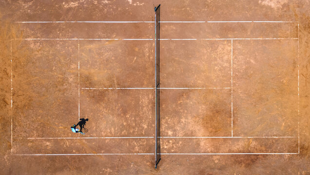 Aerial view of a lone figure with a blue bag stands on a weathered terracotta tennis court intersected by crisp white lines and a dark net, Split, Split-Dalmatia County, Croatia.