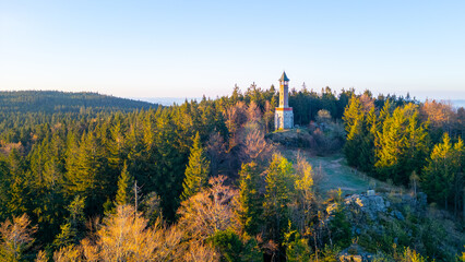 On a sunny spring morning, the Stepanka Lookout Tower stands majestically atop green hills in Korenov, surrounded by lush forests and a clear blue sky.