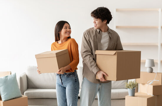 Home relocation concept. Positive young Asian couple carrying carton boxes in their owned house. Portrait of millennial tenants with belongings moving into new property