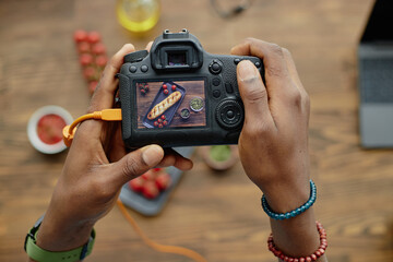Black man holding digital camera photographing food on wooden table, hands visible adjusting camera settings, image on camera screen showing plated dish with garnish and ingredients