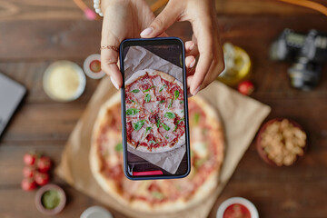 Unrecognizable woman holding smartphone photographing freshly baked pizza on wooden table, hands visible arranging phone above food, camera and ingredients in background