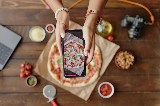 Young woman photographing freshly baked pizza with smartphone, hands holding phone above pizza on wooden table, food styling props and camera visible in background - Powered by Adobe